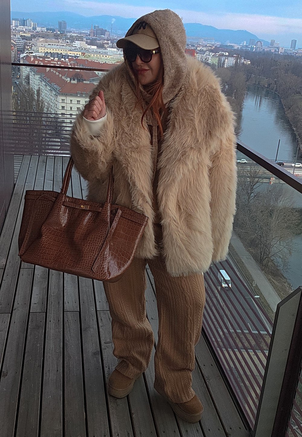 Personne portant un manteau en fourrure beige surdimensionné, un pantalon en tricot marron, des lunettes de soleil et une casquette, tenant un grand sac à main marron sur un balcon avec vue sur la ville et la rivière.