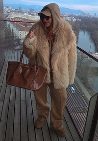 Personne portant un manteau en fourrure beige surdimensionné, un pantalon en tricot marron, des lunettes de soleil et une casquette, tenant un grand sac à main marron sur un balcon avec vue sur la ville et la rivière.