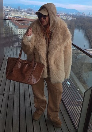 Persona con un abrigo de piel beige extragrande, pantalones de punto marrón, gafas de sol y gorra, sosteniendo un gran bolso marrón en un balcón con vista a la ciudad y al río.