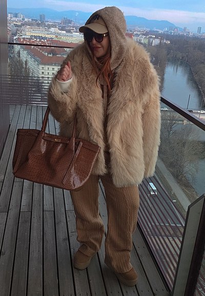 Persona con un abrigo de piel beige extragrande, pantalones de punto marrón, gafas de sol y gorra, sosteniendo un gran bolso marrón en un balcón con vista a la ciudad y al río.