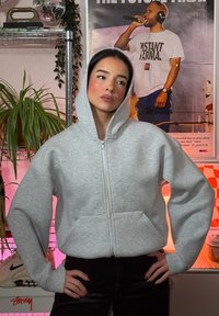 Šedá mikina na zips s priestrihom a veľkými prednými vreckami, s jemnou textúrou a nastaviteľnou kapucňou.