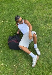 Mladý muž s copánky typu cornrow, slune�čními brýlemi, bílým tílkem, šedými kraťasy a stříbrnými teniskami odpočívá na trávě vedle černé tašky přes rameno s červeným nápisem.