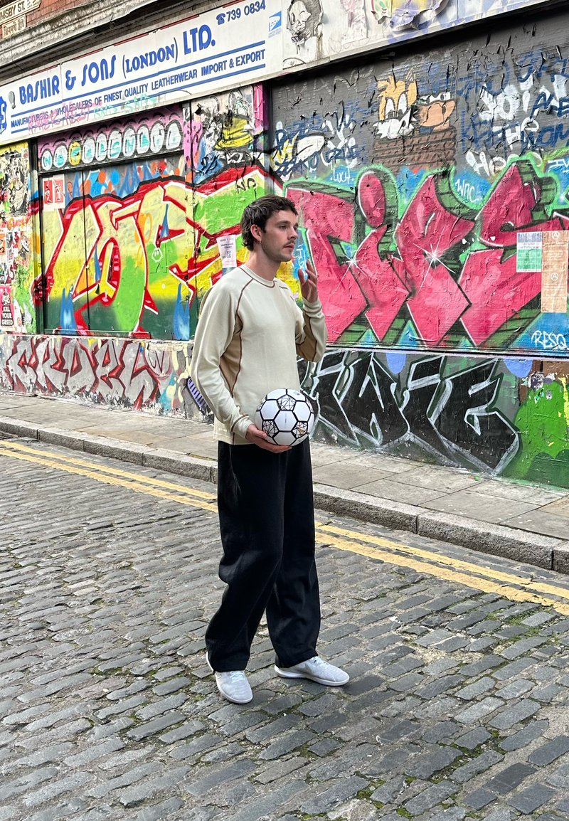 White long-sleeve shirt with beige accents, black loose pants, and white sneakers. Holding a black and white soccer ball in a graffiti backdrop.