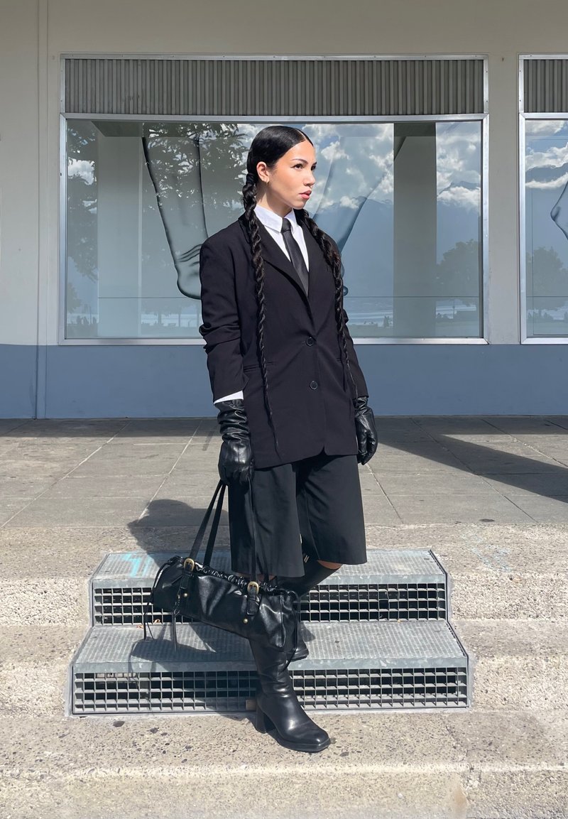 Persona con capelli lunghi intrecciati sta sulle scale indossando un blazer nero, guanti, pantaloni shorts fino al ginocchio, stivali e tiene in mano una borsa nera contro la facciata di un edificio.