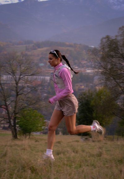 Mujer con camiseta de manga larga rosa y pantalones cortos corriendo al aire libre en una colina cubierta de césped, con árboles y montañas en el fondo.