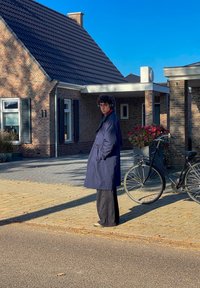 Ναυτική καμπούρα επισκιάζει έναν κόκκινο γιακά, φορεμένο πάνω από μαύρα παντελόνια. Ένα ποδήλατο στέκεται δίπλα σε έναν το brick σπίτι με λευκά περιθώρια και γλάστρες.