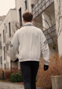 Veste bomber légère pour homme en blanc avec des accents gris, dotée de poignets élastiques et d'un col montant, associée à un pantalon de sport noir.
