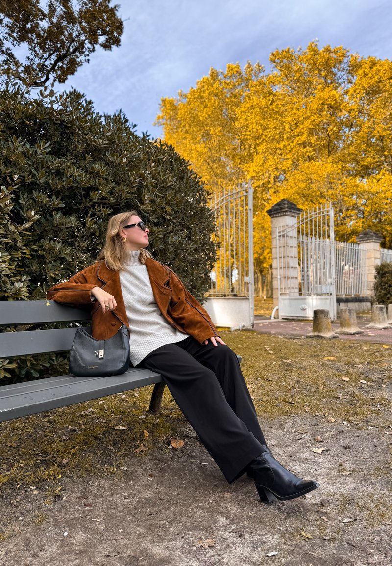 Brązowa skórzana kurtka na jasnoszarym swetrze, czarne szerokie spodnie i czarne botki na obcasie. Szara torebka leży na szarej ławce.
