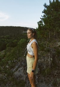 Minge sportivă cu un top alb scurt și pantaloni scurți galbeni deschis. Include un ceas, iar fundalul prezintă copaci și teren accidentat.