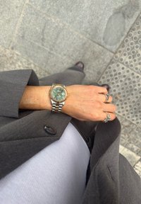 Persoană purtând un ceas de mână metalic bicolor și inele argintii, îmbrăcată într-un sacou gri și o cămașă albă, stând pe o pavăză texturată.