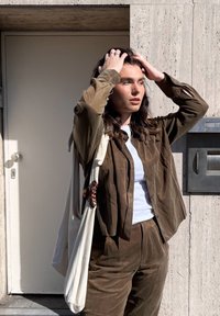 Persoană cu păr lung și închis la culoare care își aranjează părul, purtând cămașă și pantaloni maro cu un top alb, stând lângă un perete bej și o ușă în lumina soarelui.