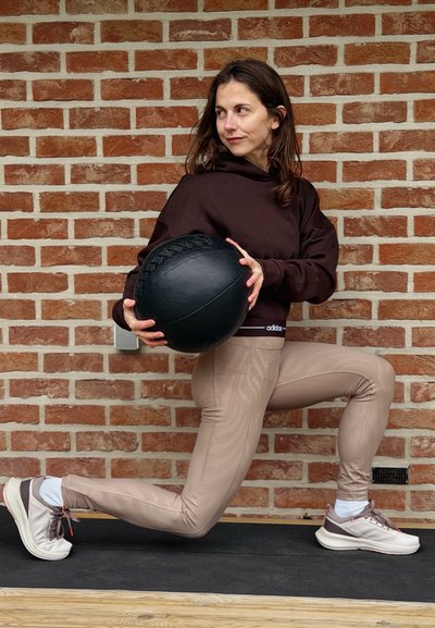 Mujer con sudadera marrón con capucha y leggins beige sosteniendo una pelota medicinal negra, realizando un ejercicio de zancada frente a una pared de ladrillos.