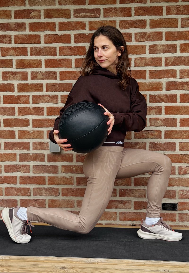 Femme en sweat à capuche marron et leggings beige tenant un ballon médicinal noir, effectuant un exercice de fente devant un mur en briques.