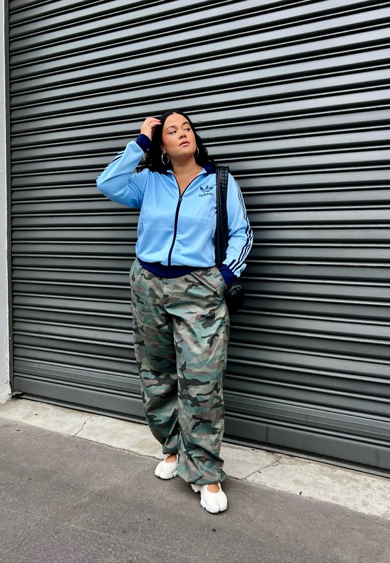 Casaco azul claro com fecho éclair, detalhes em azul escuro e logótipo da adidas, combinado com calças largas de camuflagem e ténis brancos, em pé contra um fundo riscado.