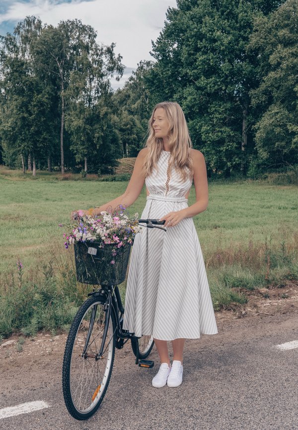 Femme en robe blanche debout à côté d'un vélo avec un panier rempli de fleurs sauvages sur une route rurale avec des arbres et de l'herbe en arrière-plan.
