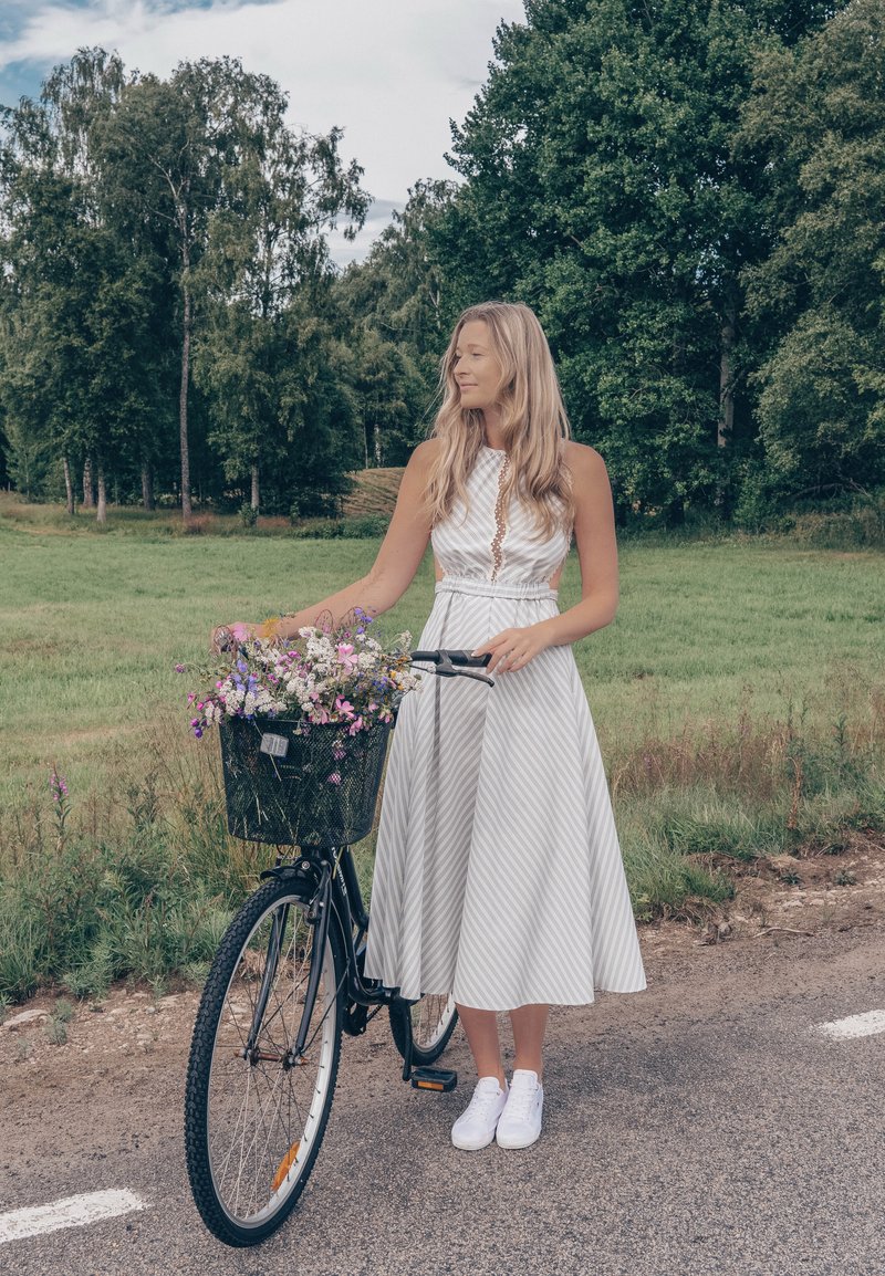 Femme en robe blanche debout à côté d'un vélo avec un panier rempli de fleurs sauvages sur une route rurale avec des arbres et de l'herbe en arrière-plan.