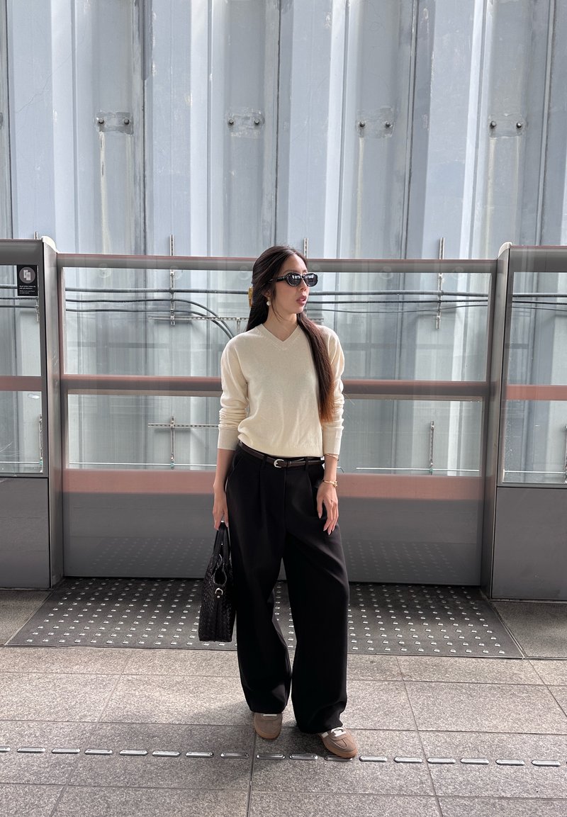 Femme portant des lunettes de soleil, un pull crème et un pantalon large noir, debout à l'intérieur tenant un sac à main noir près d'une rambarde en verre.