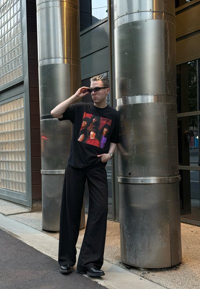 Camiseta gráfica negra con impresiones coloridas de rostros, combinada con pantalones holgados negros y zapatos negros. En el fondo hay pilares de metal y cristal.