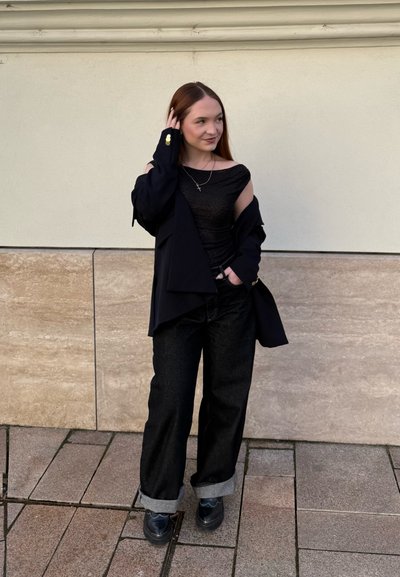Mujer de pie en la acera con pantalones negros con puños, top negro sin hombros y chaqueta negra, tocándose el cabello y sonriendo ligeramente.