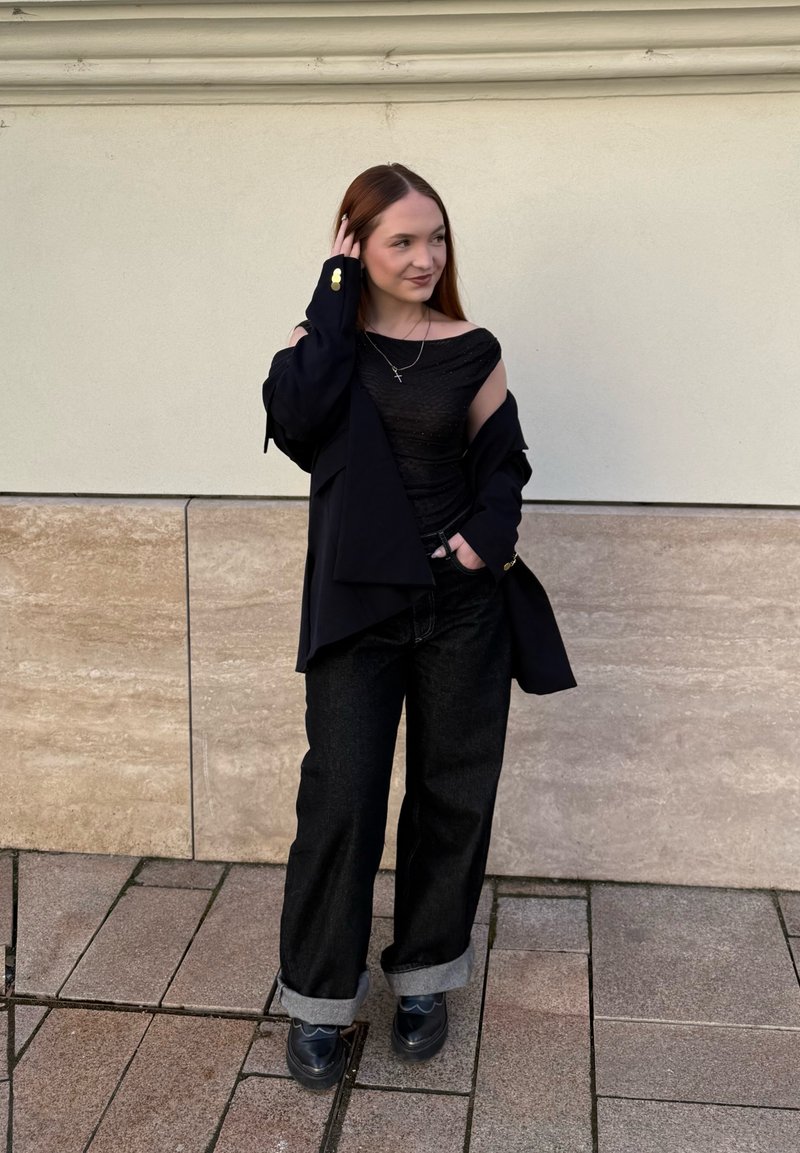 Femme debout sur le trottoir en pantalon noir avec poignets, top noir épaules dénudées et veste noire, touchant ses cheveux et souriant légèrement.