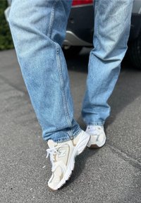 Zapatillas deportivas blancas y beige con parte superior de malla y sintético, combinadas con pantalones vaqueros de denim azul claro, de pie sobre un fondo de pavimento gris.