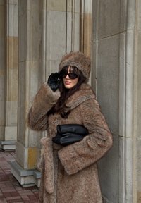 Manteau marron en fausse fourrure avec un large col et ceinture, assorti d'un chapeau en fourrure. Le modèle tient une pochette noire et porte des gants noirs ainsi que des lunettes de soleil.
