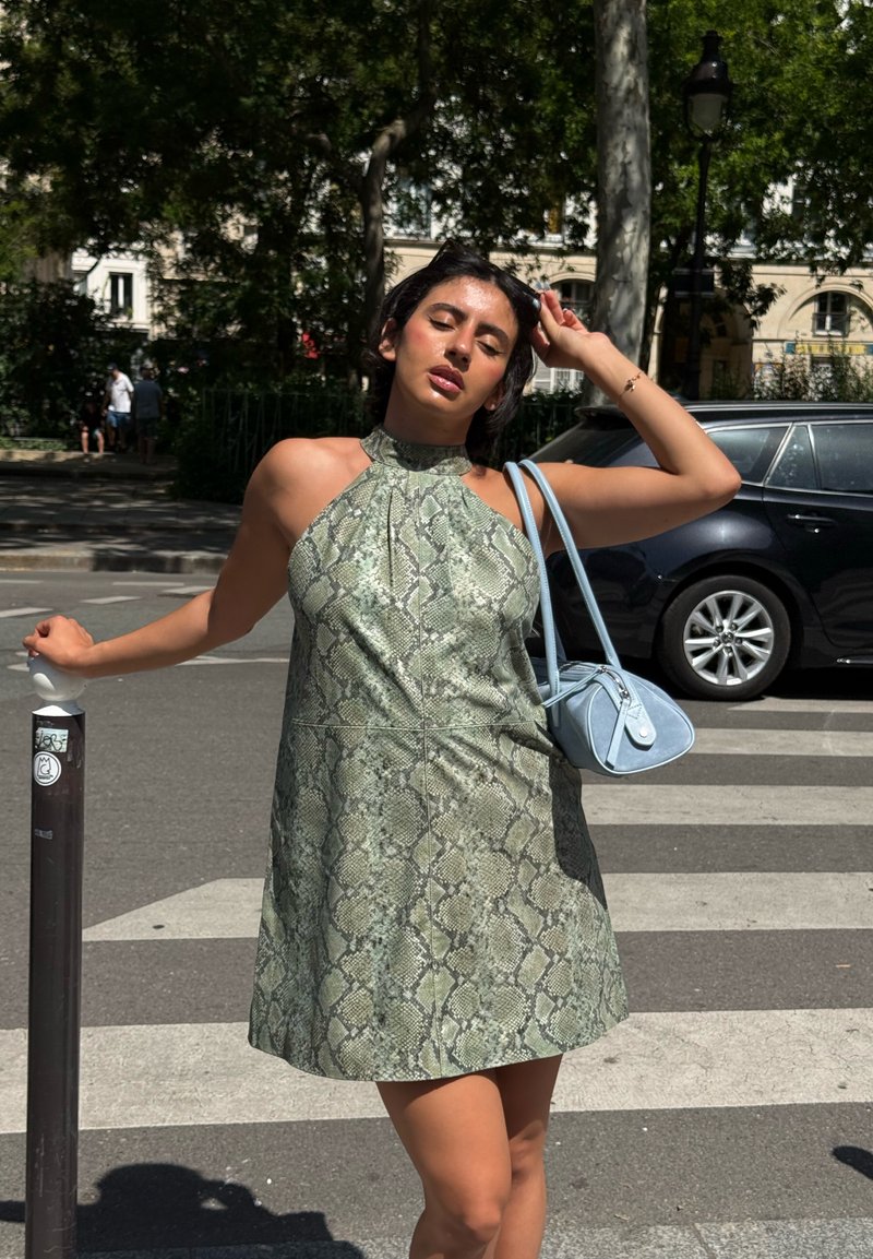Femme en robe vert imprimé peau de serpent se tient sur un passage piéton en ville, tenant un sac à main bleu clair et touchant ses cheveux les yeux fermés.