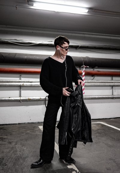 Suéter negro de punto de manga larga, pantalones anchos negros, sosteniendo una chaqueta de cuero negra. Los accesorios incluyen auriculares blancos y gafas de sol.