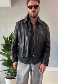 Giacca di pelle nera con zip frontale, collo appuntito e vestibilità sagomata, indossata sopra una maglietta nera e abbinata a pantaloni grigio chiaro.