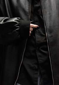 Veste noire zippée avec une texture en cuir lisse et des poignets côtelés, superposée à une chemise sombre boutonnée, présentant un design élégant.