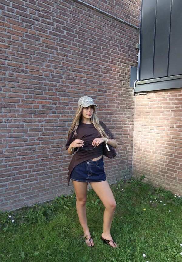 Junge Frau mit Mütze, dunklem Hemd, Jeansrock und Sandalen steht am Tag auf Gras neben einer Ziegelwand, hält Sonnenbrille und Tasche.