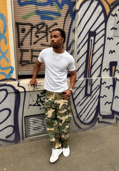 Hombre con camiseta blanca, pantalones de camuflaje y zapatillas blancas está apoyado en una pared cubierta de grafitis con una mano en el bolsillo y la otra apoyada en un saliente.