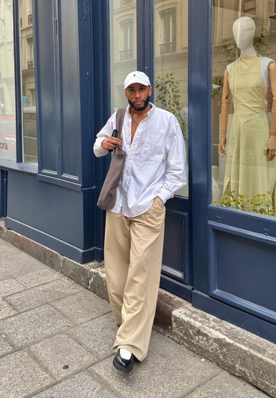 Camisa blanca de botones, pantalones beige anchos y zapatos negros. Un bolso gris cuelga del hombro. Hay un maniquí con un vestido amarillo cerca.