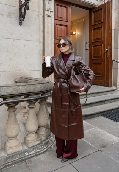 Trench de cuero marrón con solapas anchas y cintura ceñida, combinado con pantalones burdeos y tacones a juego; sosteniendo una taza de café.