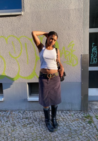 Top blanco sin mangas, falda de pana morada, cinturón verde con una hebilla circular. Botas negras hasta la rodilla. De pie contra una pared gris con grafitis.