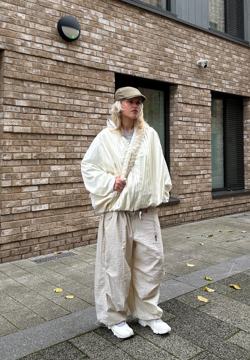 Witte, oversized jas met geribbelde textuur en verlaagde schouders, gecombineerd met losse beige broek. Gestyled met een lichtgroene pet en witte sneakers.
