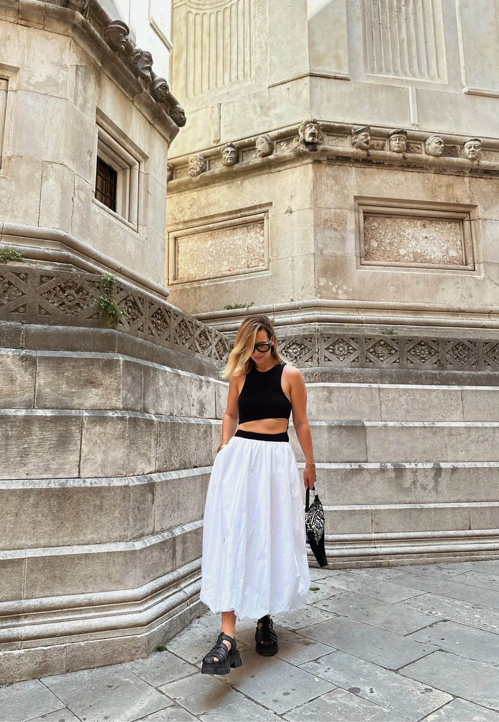 Top cropped preto, saia branca fluida e sandálias pretas robustas. Os acessórios incluem uma bolsa com padrão. Muro de pedra com detalhes ornamentais ao fundo.