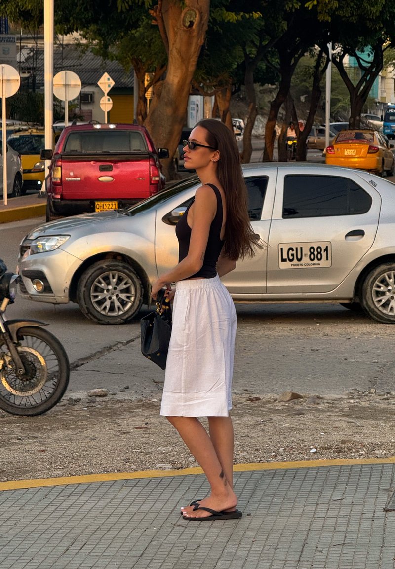 Schwarzes Tanktop mit dünnen Trägern, weiße knielange Rock, schwarze Handtasche, Flip-Flops, langes braunes Haar, städtische Straße mit Fahrzeugen.