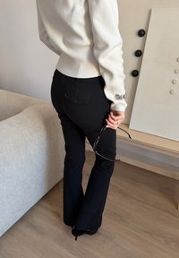 Personne portant un pantalon noir et un pull blanc, tenant des lunettes à monture noire, debout à côté d'un canapé beige et d'un panneau mural décoratif.