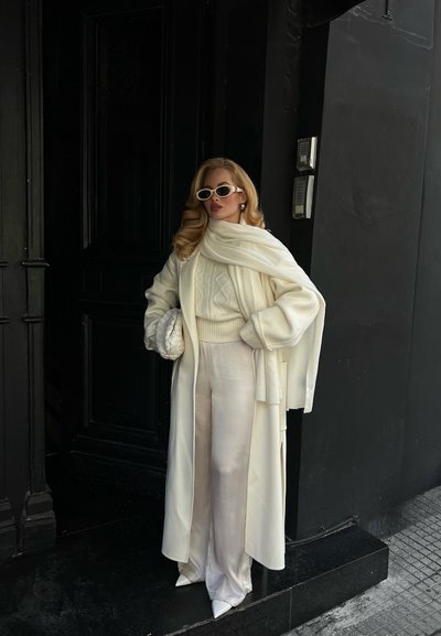 Mujer vestida con un conjunto completamente blanco que incluye un abrigo largo, bufanda, suéter, pantalones anchos y gafas de sol blancas, sosteniendo un bolso tejido, de pie junto a una puerta oscura.
