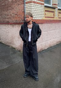 Svart bomberjakke over en hvit singlet, løse svarte bukser og robuste svarte støvler. Bølgete hår, briller og et sølvkjede.