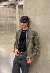 Jachetă din piele maro cu fermoar, tricou negru, blugi de denim închis. Accesoriile includ un colier din lanț argintiu și cercei mici cu cerc.