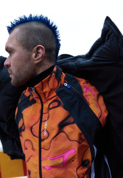 Chaqueta multicolor con un diseño en naranja y negro, con patrones rosas, cuello alto y cremalleras. Tela texturizada con un corte deportivo.