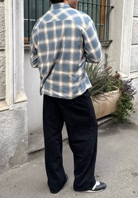 Light blue check shirt with white accents, black wide-leg trousers, and black shoes with white detailing, standing on a paved surface.
