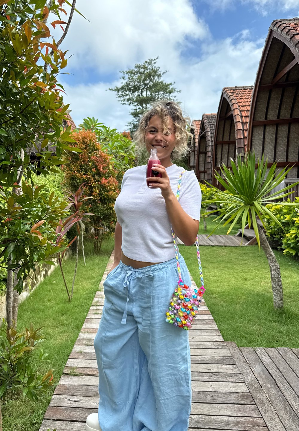 Lyseblå linned bukser med vid ben, hvid ribbet crop top og en farverig perle taske. Holder en klar flaske med mørk væske. Grønne planter i baggrunden.