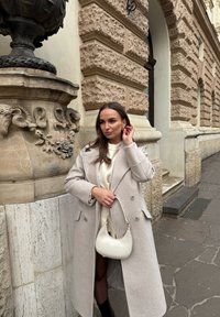 Abrigo de mezcla de lana gris claro con solapas oversized, combinado con un suéter crema y botas negras hasta la rodilla. Lleva un pequeño bolso blanco.
