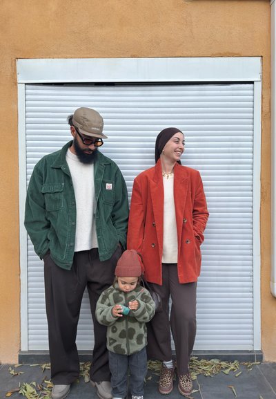 Hombre con chaqueta verde y gorra, mujer con blazer naranja sonriendo, niño con chaqueta estampada y gorro jugando con un juguete, de pie frente a una persiana blanca cerrada.