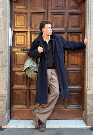 Joven con abrigo largo oscuro y pantalones anchos marrones apoyado contra una puerta de madera ornamentada, llevando un bolso verde de lona al hombro.