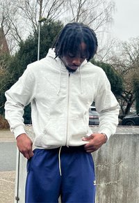Homme aux dreadlocks portant un sweat à capuche blanc zippé et un pantalon de survêtement Adidas bleu ajuste ses vêtements en plein air, près d'arbres et de voitures garées.