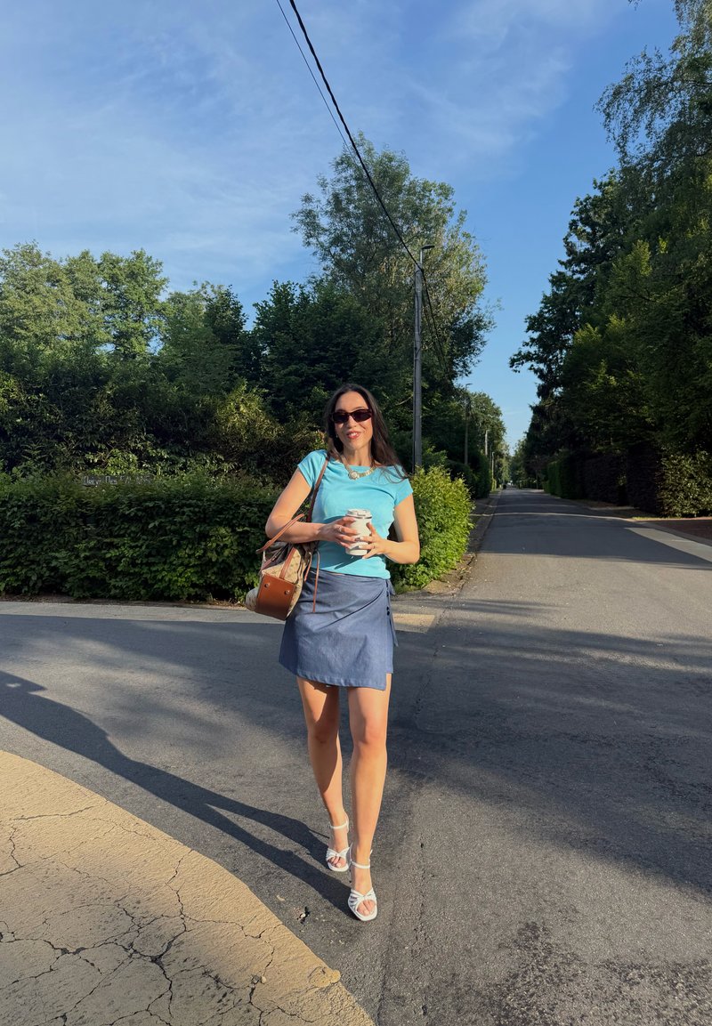 Femme portant des lunettes de soleil, un haut bleu et une jupe marche sur une route ensoleillée en tenant une tasse de café, avec des arbres et des buissons en arrière-plan.